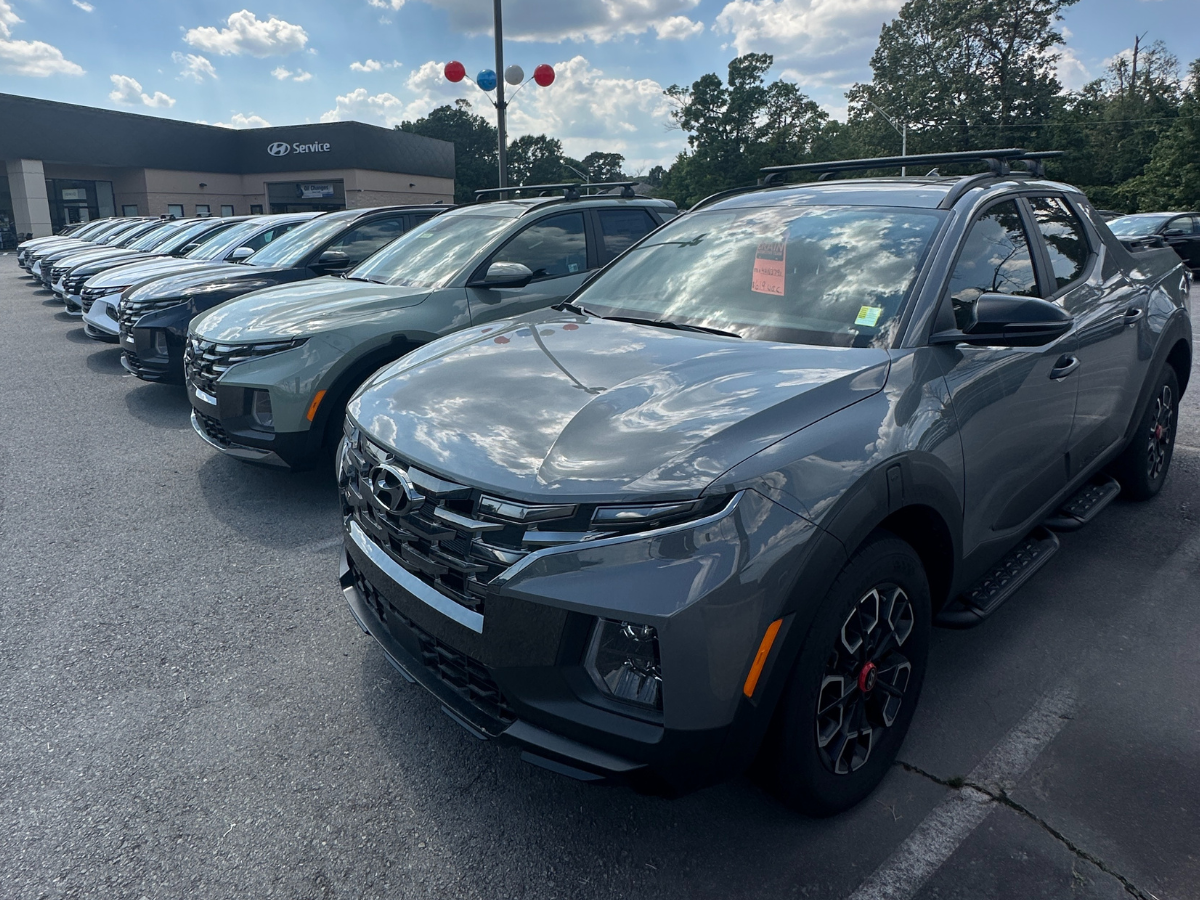 A front view of multiple new 2024 Hyundai models, including the Santa Cruz, lined up at Crain Hyundai of North Little Rock with blue skies and balloons in the background.