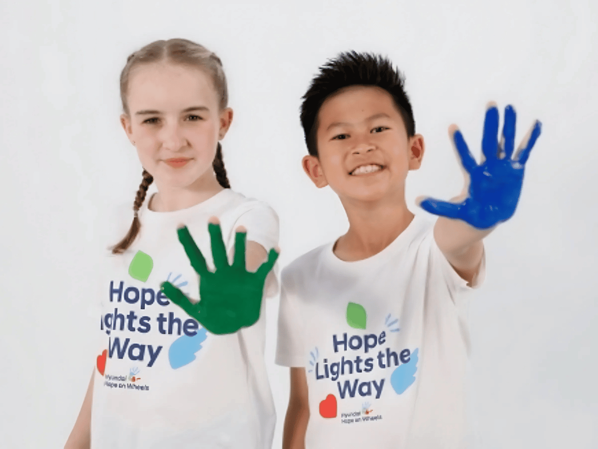 Two children wearing “Hope Lights the Way” Hyundai Hope On Wheels shirts proudly show their green and blue painted handprints toward the camera.