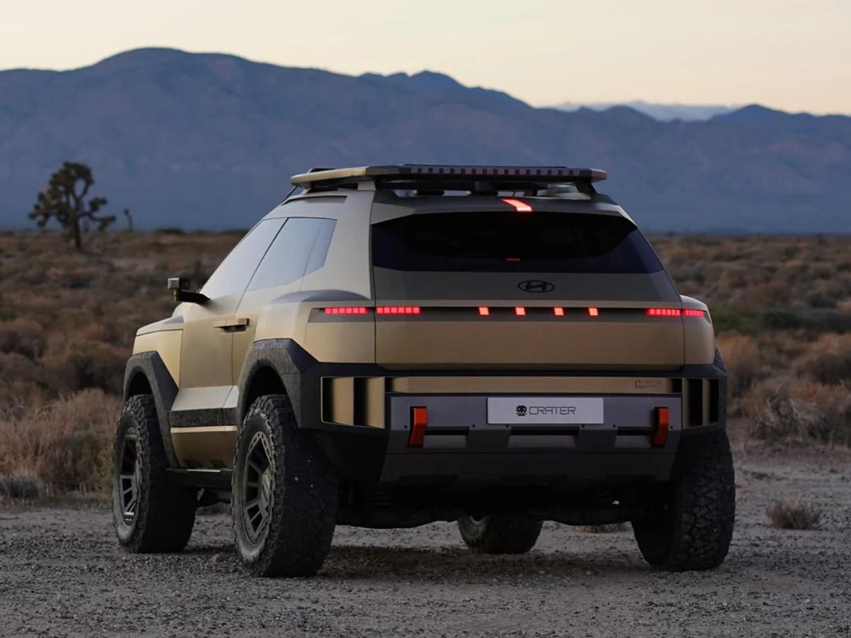 A view from the back of the gold Hyundai CRATER Concept EV as it sits in a desert. The image shows the rugged rear bumper, orange tow hooks, wide off-road tires, and the unique red pixel lighting across the tailgate.
